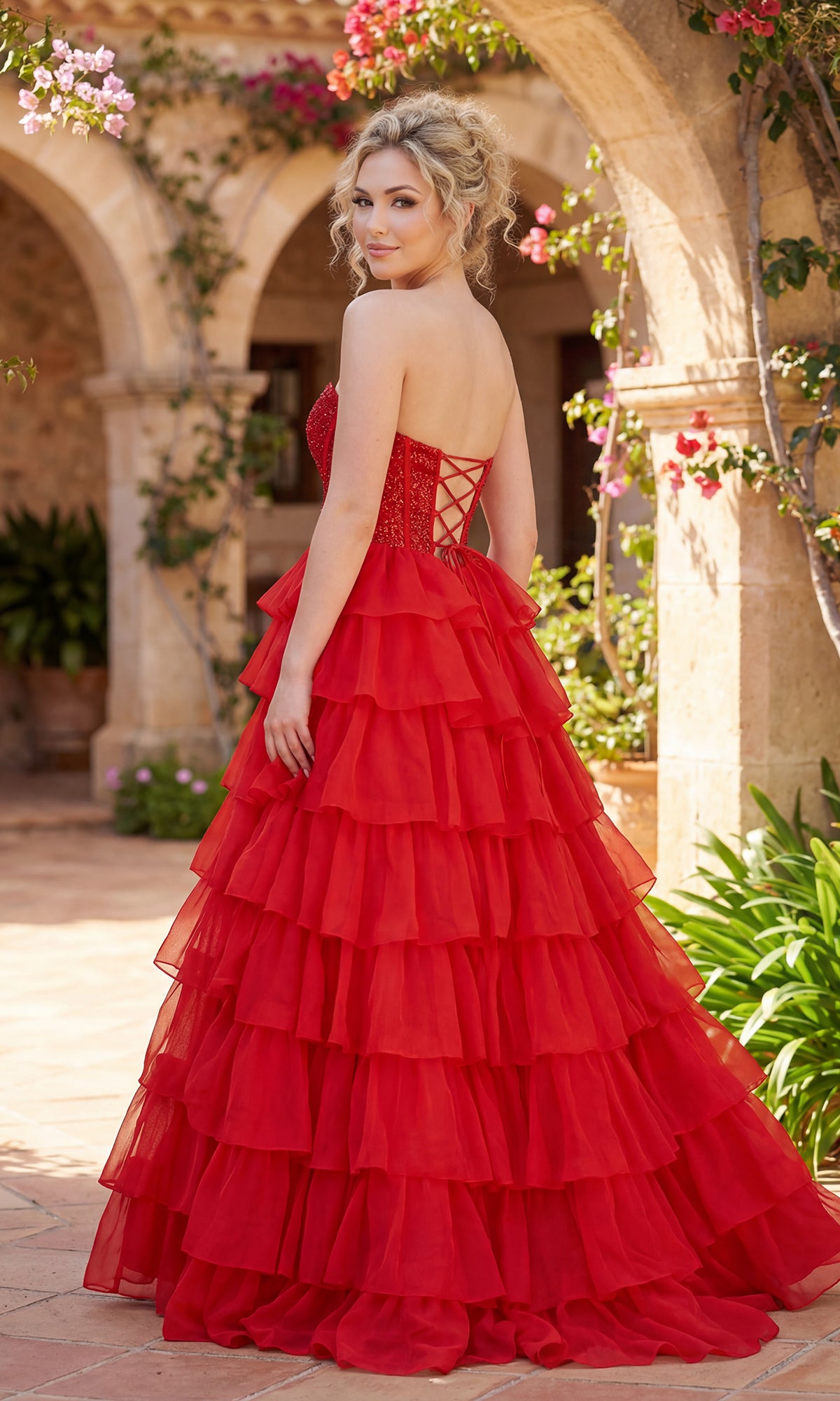 Woman in the PromGirl Exclusive 250204, a long ruffled prom dress with a beaded corset bodice, stands outdoors on a stone patio with arches and flowering plants, looking over her shoulder.